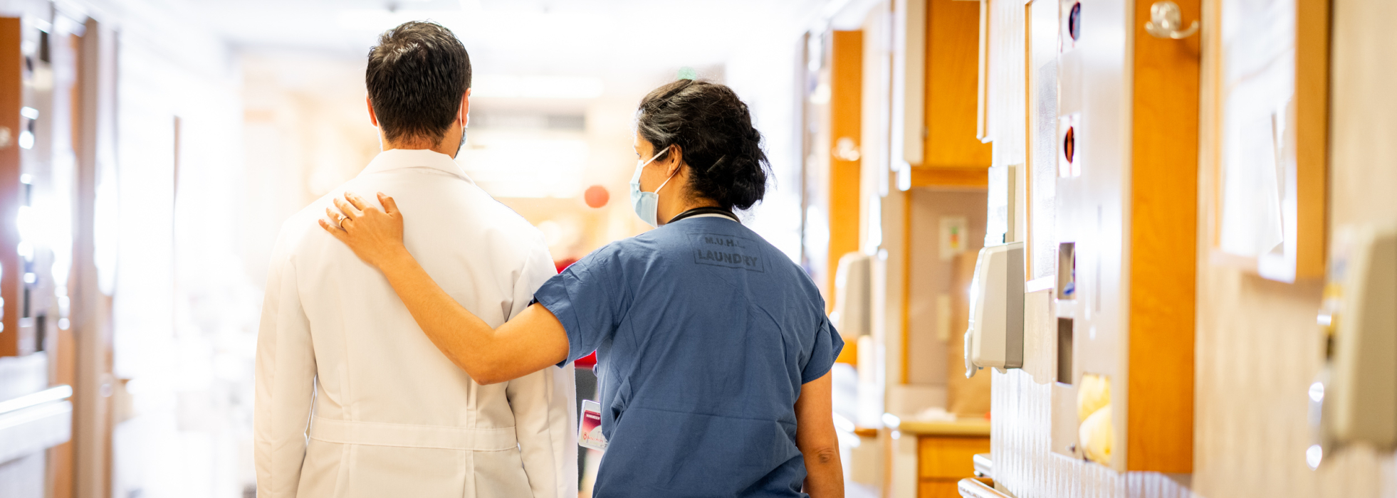 A female clinician in blue scrubs touches the shoulder of a male colleauge in a white coat while walking in the hospital hallway