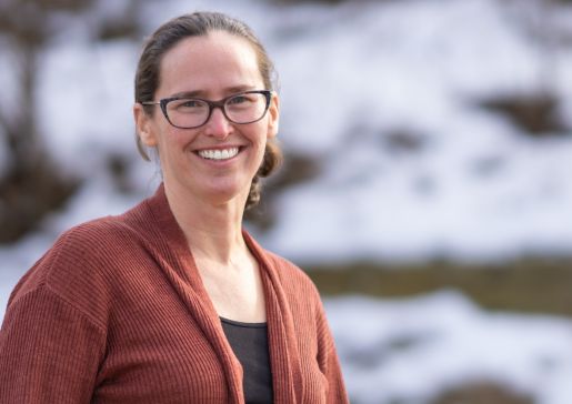 Dr. Meghan Brennan stands outside wearing dark-rimmed glasses and an orange sweater
