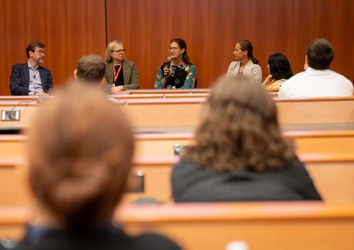 A panel of speakers sits at the front of the room, with people sitting in the audience out of focus in the foreground